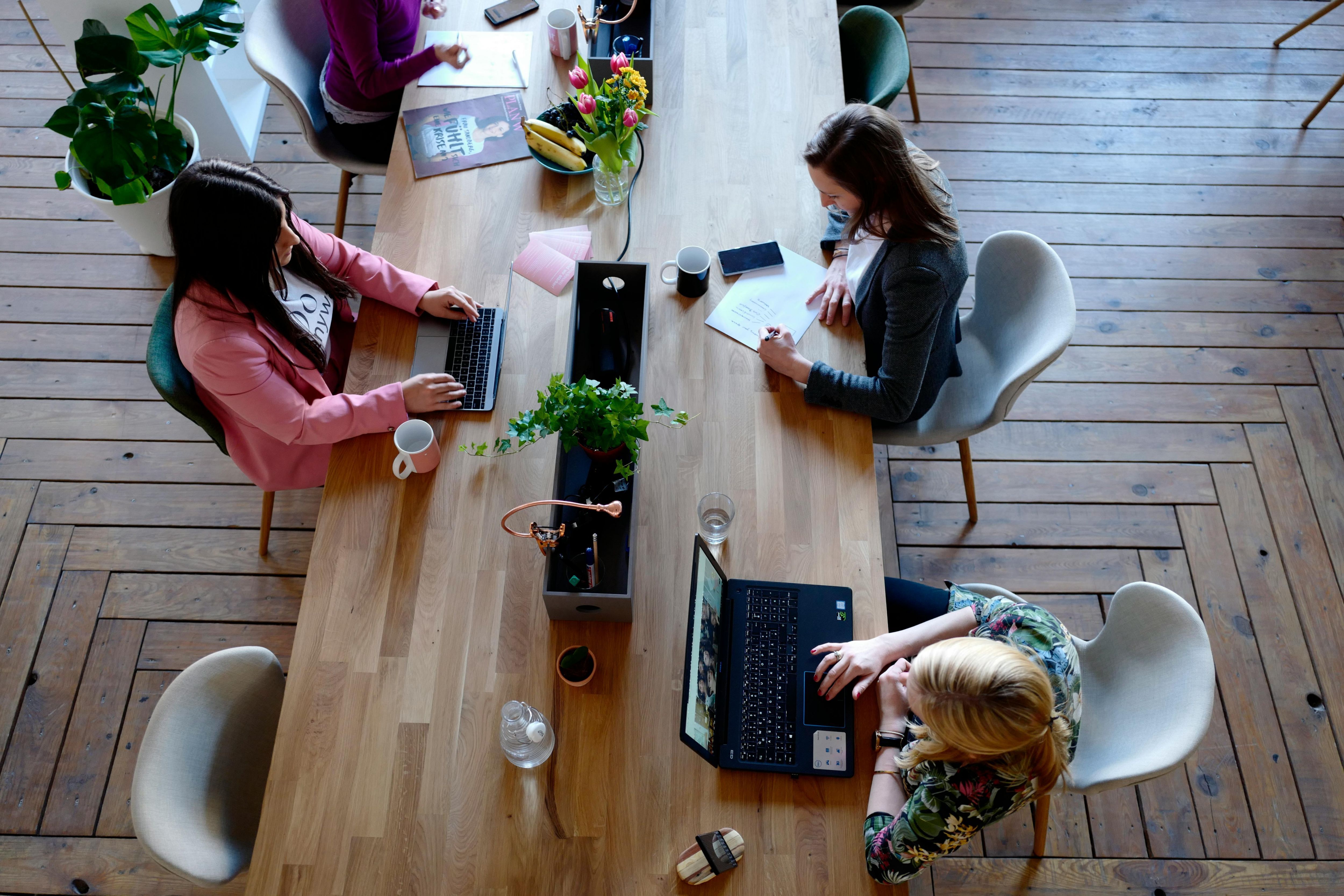 Team von Mitarbeiterinnen arbeitet gemeinsam an einem großen Tisch, entwickelt Konzepte für betriebliches Gesundheitsmanagement und optimiert Unternehmensprozesse